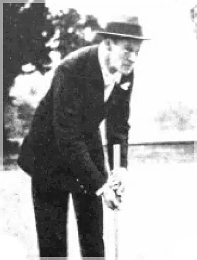 A man in a suit and hat bends forward holding a croquet mallet on a grassy field, with trees visible in the blurred background.