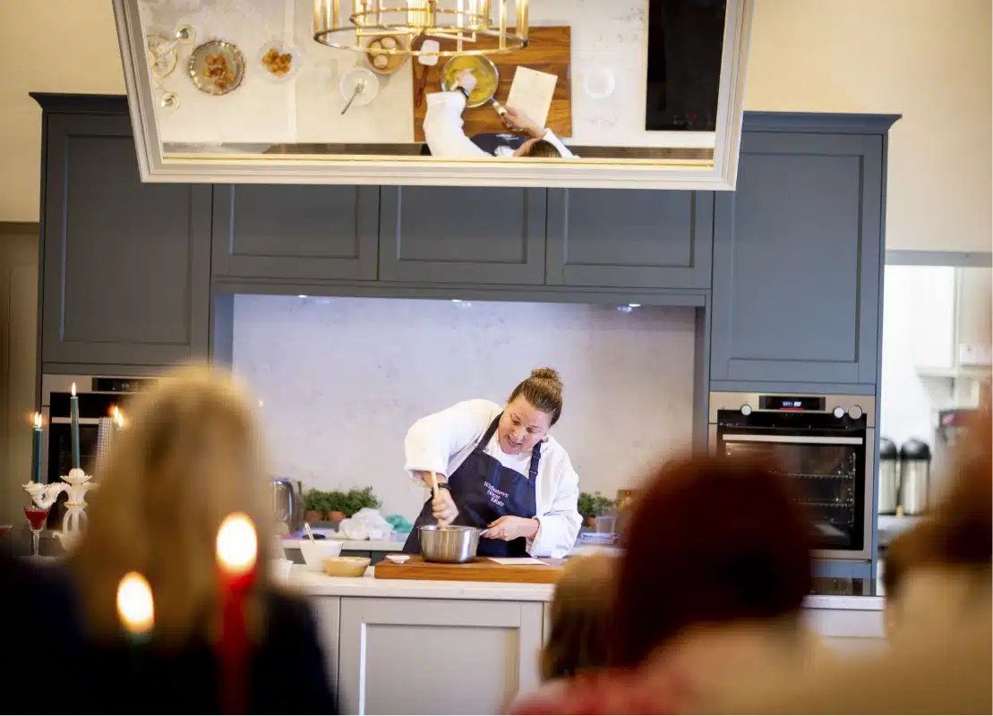 At a cooking demonstration near Malahide Castle, a chef in a white apron and blue shirt artfully stirs ingredients in a metal bowl on the counter, his movements mirrored above. In the foreground, a blurred audience watches intently.