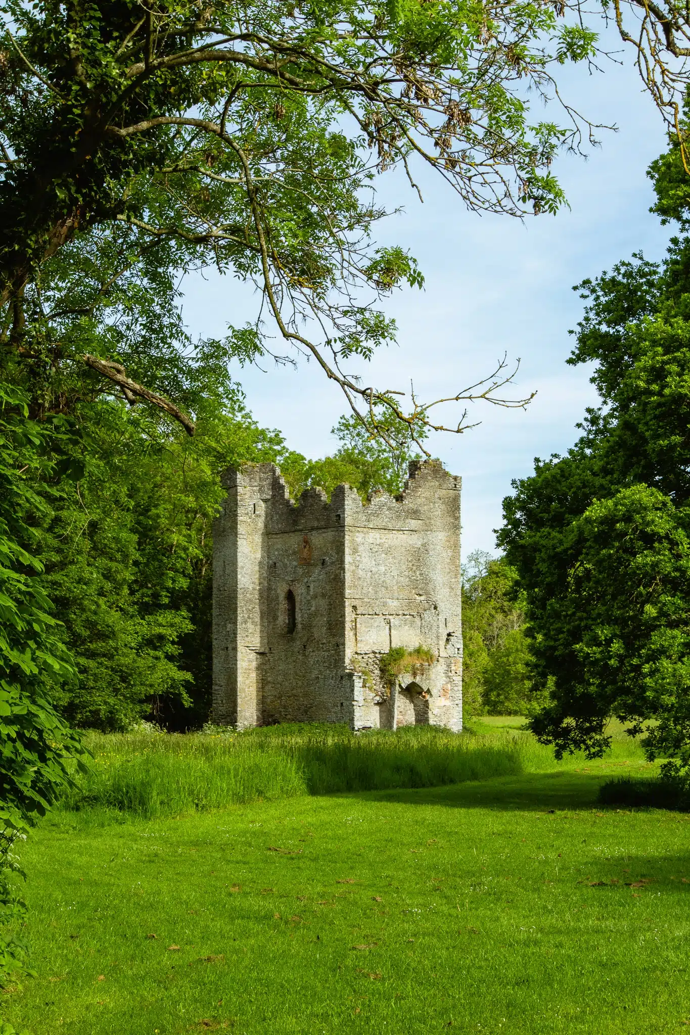 A small, old stone tower with arched windows stands in a lush, green clearing near Malahide Castle, surrounded by trees and tall grass under a bright, partly cloudy sky.