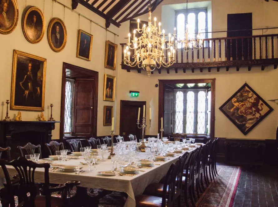 A grand dining room at Malahide Castle Properties features a long table set for a meal, adorned with white tablecloth and elegant dinnerware. Portraits and paintings decorate the walls, a large chandelier hangs from the ceiling, while natural light streams through tall windows.