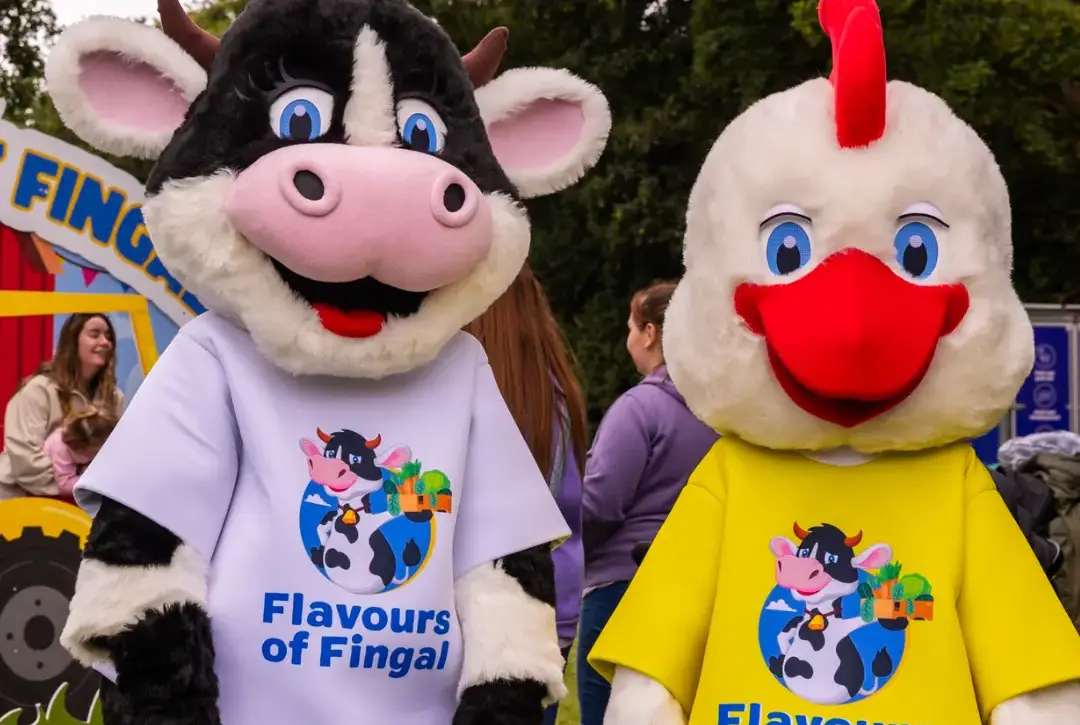 Two people in cow and chicken costumes, wearing “Flavours of Fingal” shirts, stand outdoors at an event with people and trees in the background.