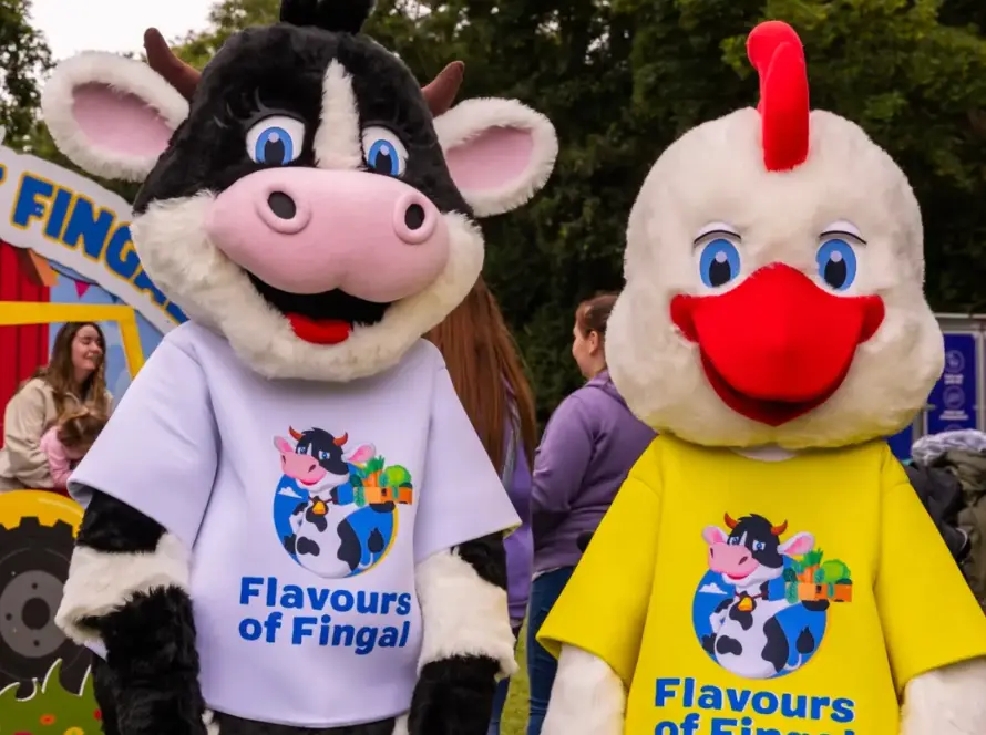 Two people in cow and chicken costumes, wearing “Flavours of Fingal” shirts, stand outdoors at an event with people and trees in the background.