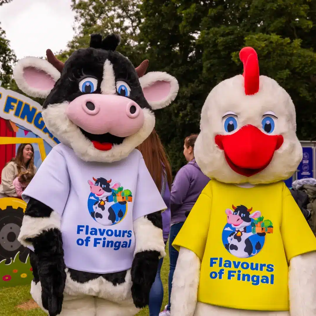 Two people in cow and chicken costumes, wearing “Flavours of Fingal” shirts, stand outdoors at an event with people and trees in the background.