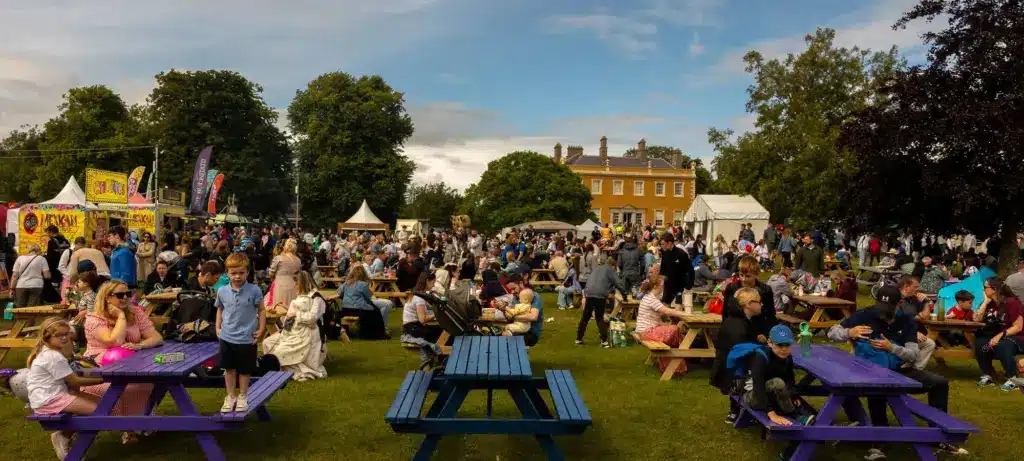 A large crowd enjoys an outdoor festival on a grassy lawn with picnic tables, food stalls, and a historic building in the background under a partly cloudy sky.