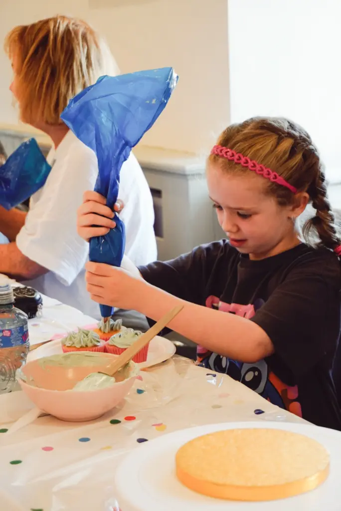 A young girl with braided hair decorates a cupcake using a blue piping bag, concentrating intently. She sits at a table covered with baking supplies, enjoying her time at the Little Baker Cupcake Classes.