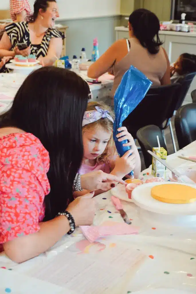 A woman helps a young girl at the table during Little Baker Cupcake Classes. The focused girl holds a blue piping bag, while baking supplies cover the table and others work in the background.