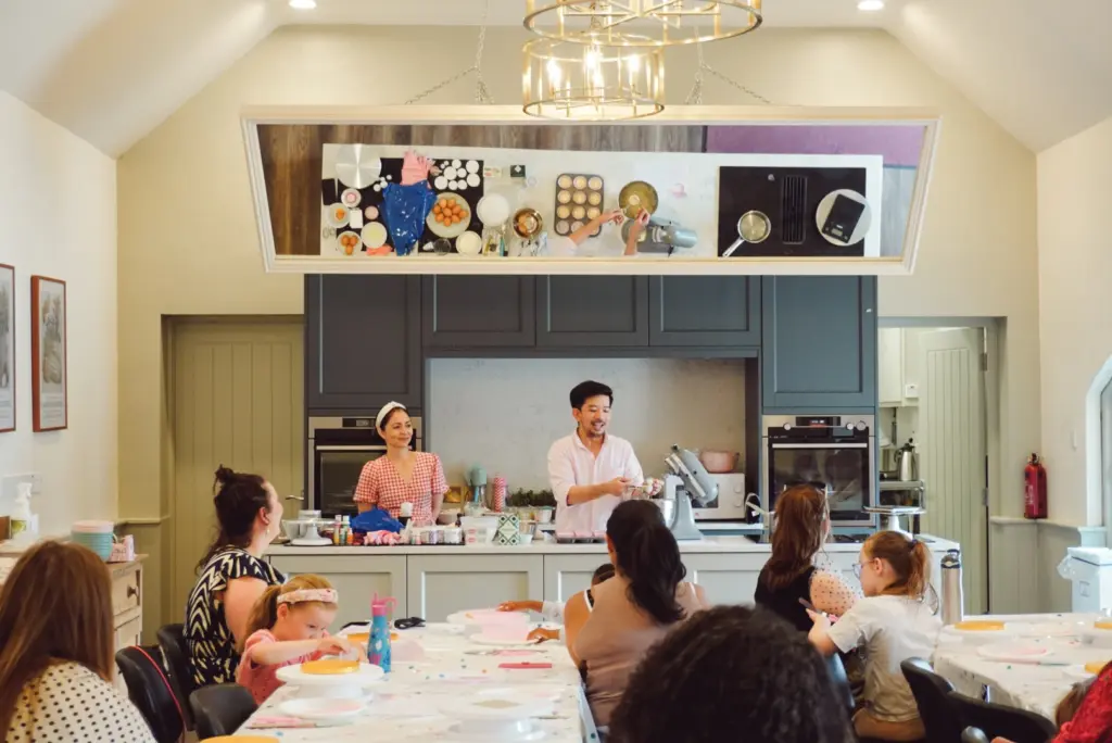 A man and woman stand behind a kitchen counter leading a lively Little Baker / Big Baker Cupcake Class, with students seated at tables watching. A large mirror above reflects the countertop and baking ingredients in the well-lit room.