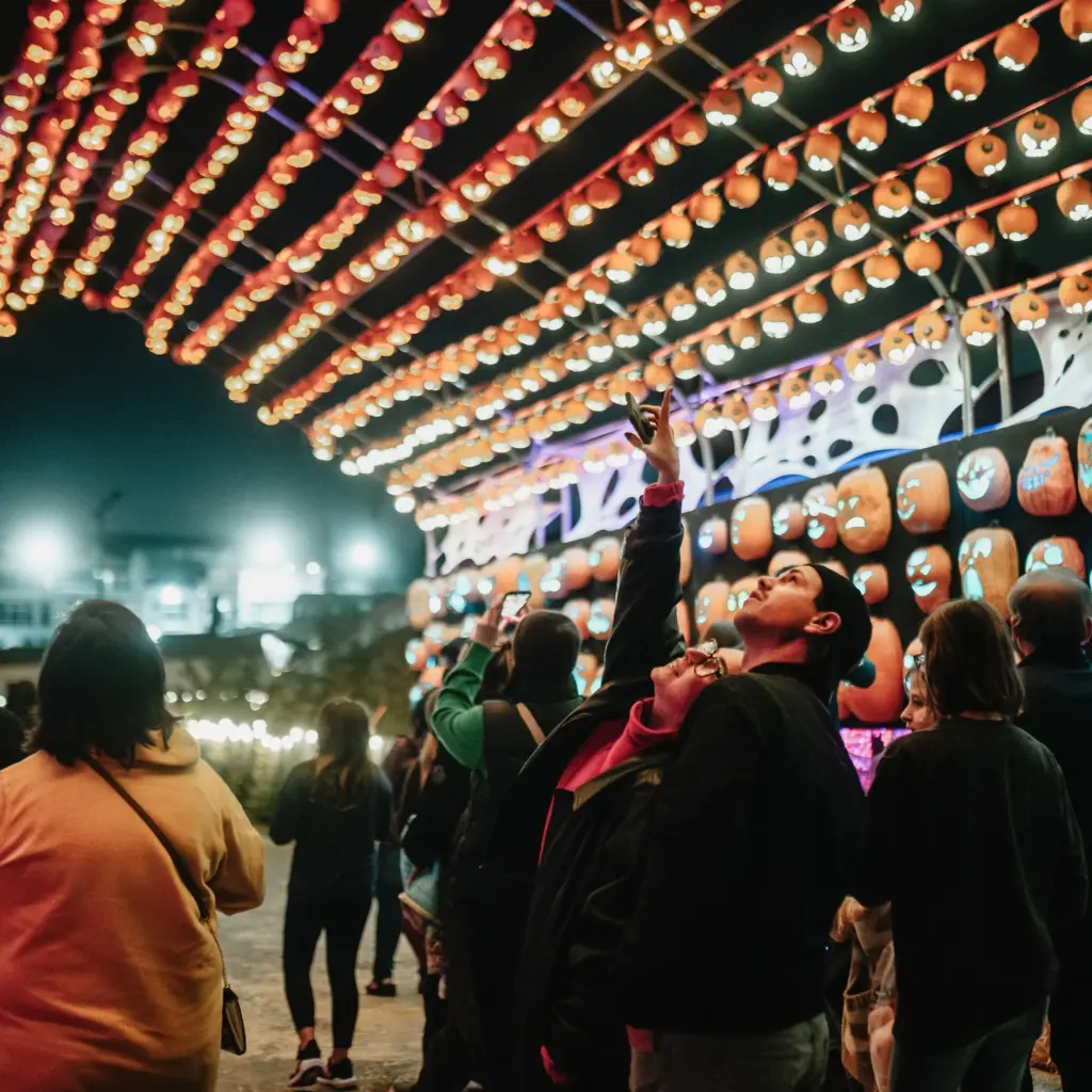 A group of people, some taking photos, walk under a canopy of glowing orange lanterns at night during Pumpkins After Dark: Ireland's Spectacular Halloween Experience, where vibrant lights create a truly festive and magical atmosphere.