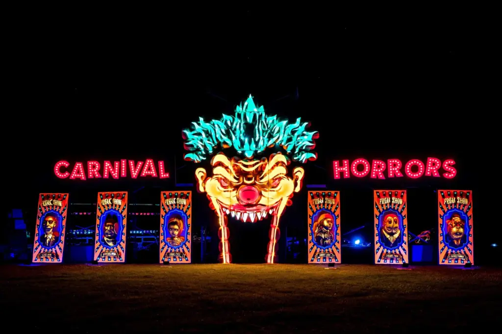 A brightly lit carnival entrance at night features a large, menacing clown face with glowing eyes and sharp teeth between neon signs reading "Carnival" and "Horrors," with creepy clown posters and flyers for Little Baker Cupcake Classes on either side.