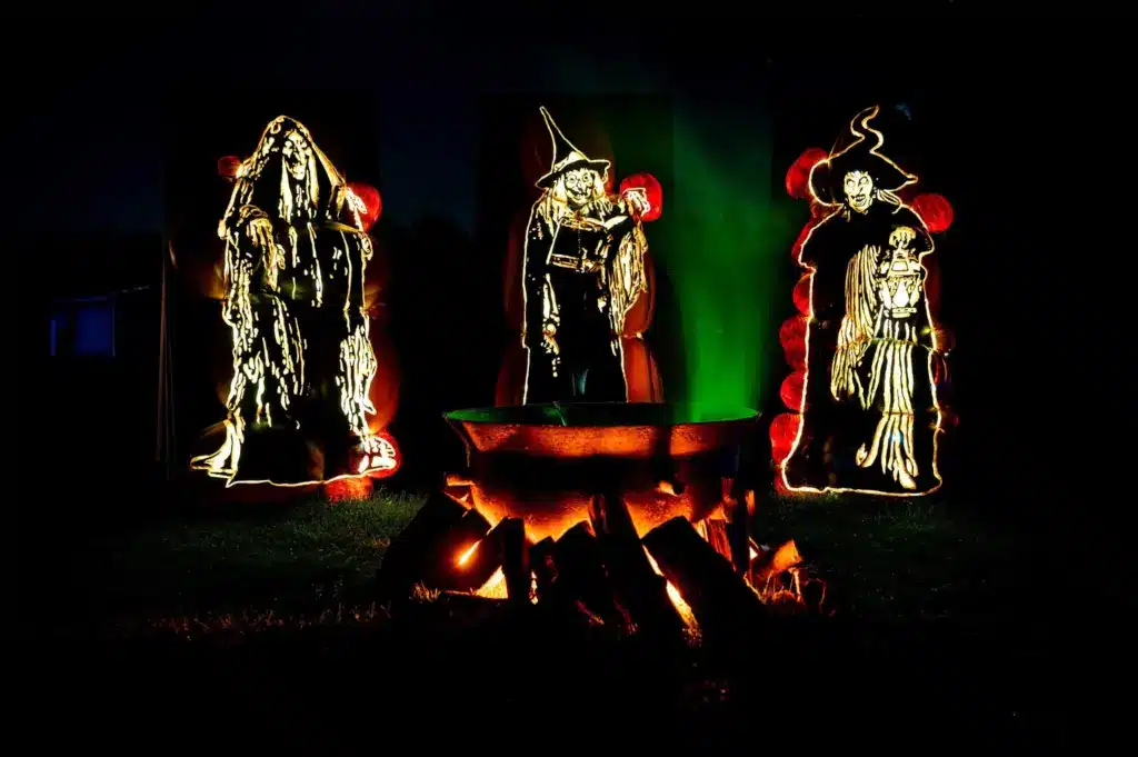 Three glowing, carved pumpkin witches stand behind a cauldron surrounded by burning logs at night, with green mist rising—a spooky Halloween scene perfect for inspiring Little Baker or Big Baker Cupcake Classes.