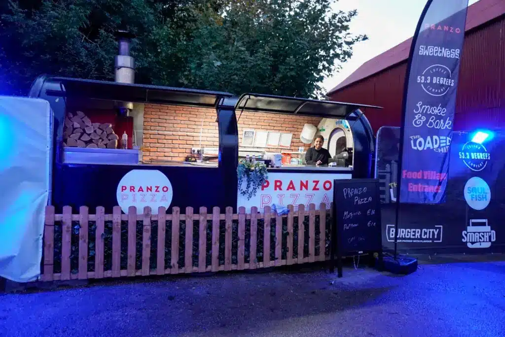 Outdoor pizza food truck "Pranzo Pizza" features a brick oven, wood logs, and two staff. A small wooden fence, menu board, and banners—including one for Pumpkins After Dark: Ireland's Spectacular Halloween Experience—surround the truck.
