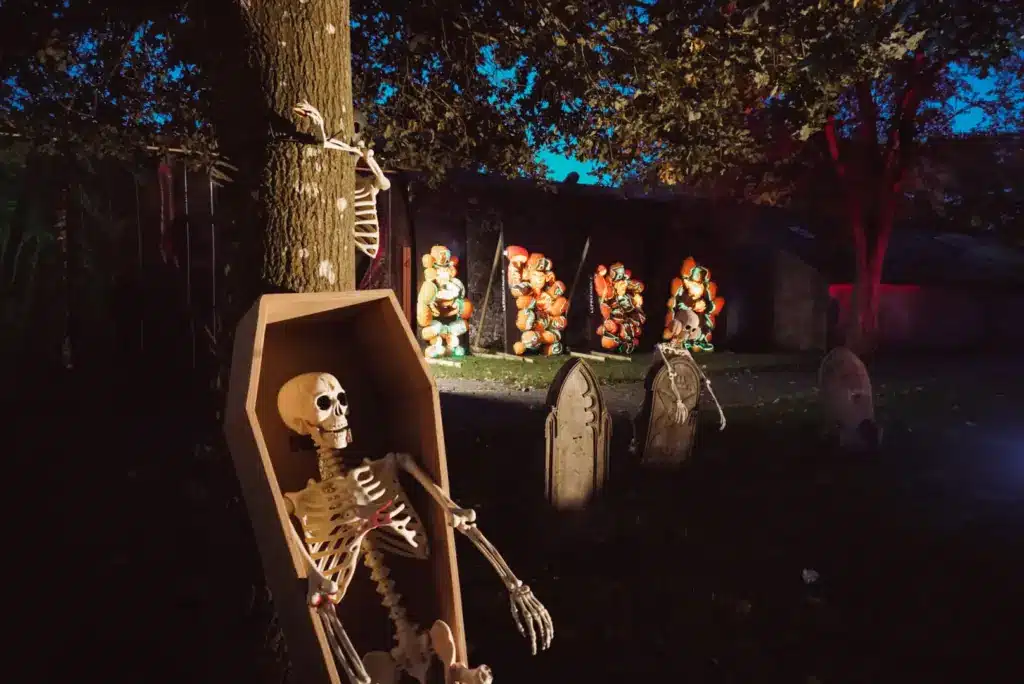 A skeleton sits upright in a coffin leaning against a tree in a graveyard at night, surrounded by tombstones and glowing pumpkin decorations—capturing the eerie magic of Pumpkins After Dark: Ireland's Spectacular Halloween Experience.