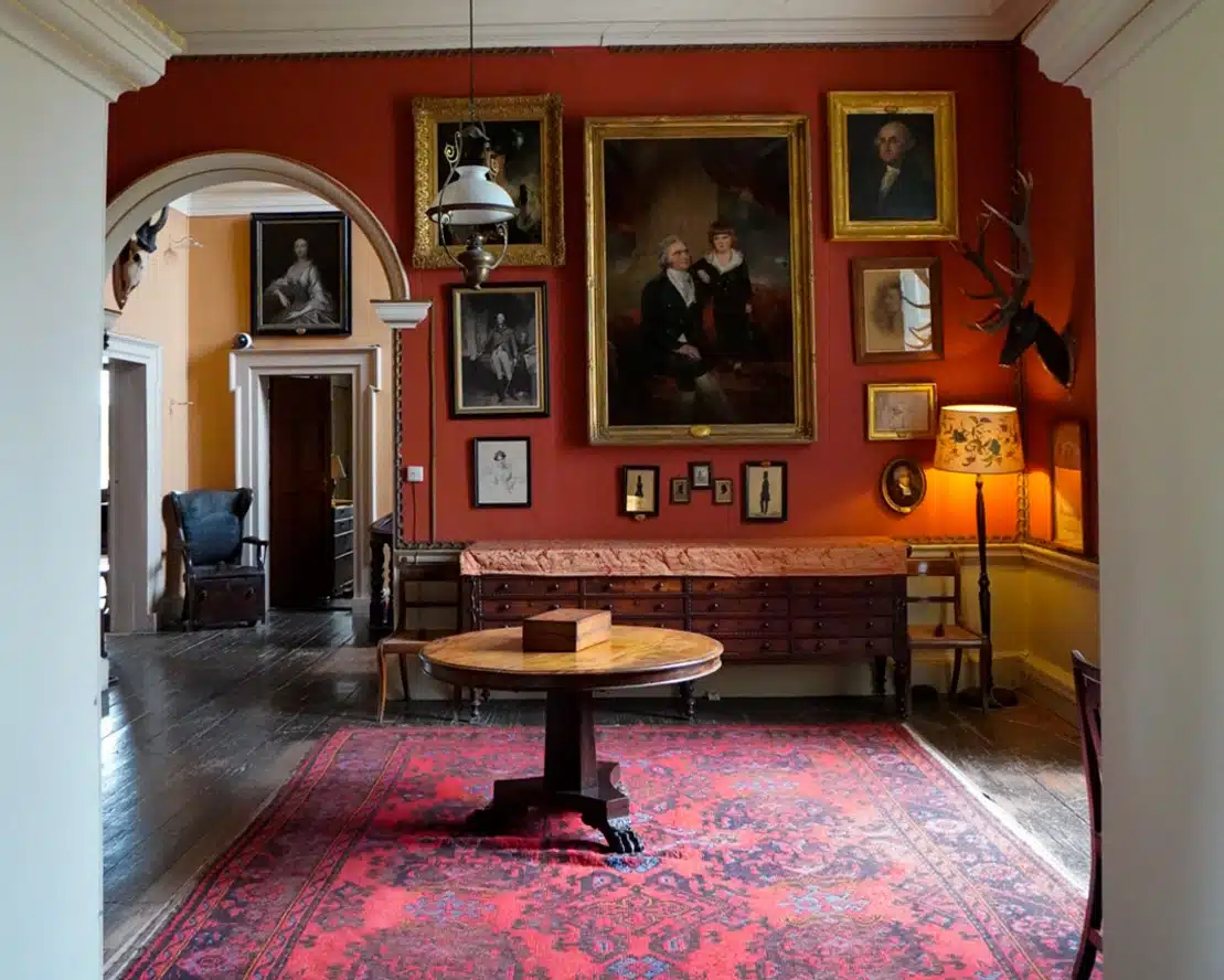 A richly decorated room with red walls, perfect for a Georgian House Tour Dublin, features multiple framed portraits, a round wooden table on a red patterned rug, antlers on the wall, and elegant wooden furniture under an archway.