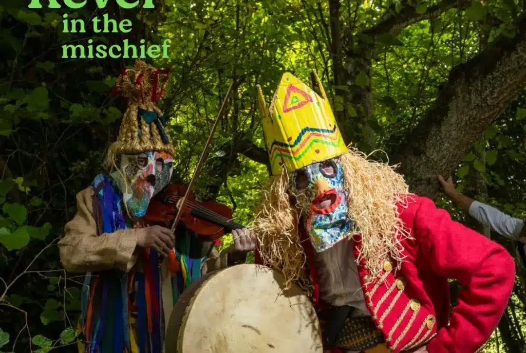 Two people in colorful, elaborate costumes and masks stand in a green forest. One plays a violin, the other holds a drum. Text reads "Revel in the mischief"—a scene reminiscent of festivities at Newbridge House and Farm