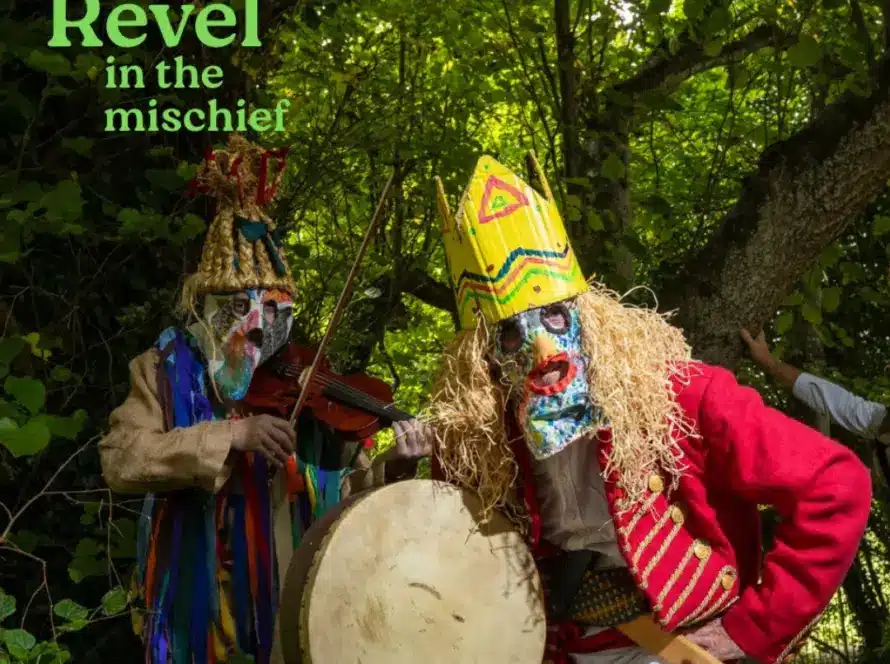 Two people in colorful, elaborate costumes and masks stand in a green forest. One plays a violin, the other holds a drum. Text reads "Revel in the mischief"—a scene reminiscent of festivities at Newbridge House and Farm