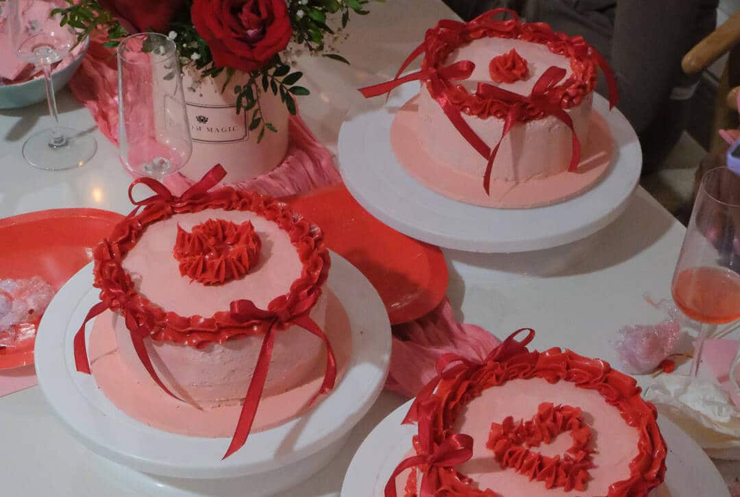 Three round pink cakes with red icing and ribbon decorations sit on white cake stands, surrounded by a bouquet of red roses, wine glasses, red plates, and pink rose petals—a scene perfect for an ABBA by Moonlight celebration.