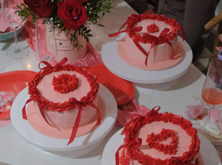 Three round pink cakes with red icing and ribbon decorations sit on white cake stands, surrounded by a bouquet of red roses, wine glasses, red plates, and pink rose petals—a scene perfect for an ABBA by Moonlight celebration.