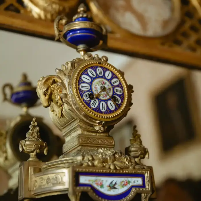 An ornate antique clock with an intricate gold frame and blue enamel detailing, reminiscent of the timeless elegance found in Malahide Castle. Roman numerals accentuate the clock face, which sits atop a decorative base set in a vintage interior with a blurred background.
